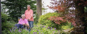 im Garten: eine Frau schiebt eine ander eim Rollstuhl an schönen Blumenbeeten vorbei
