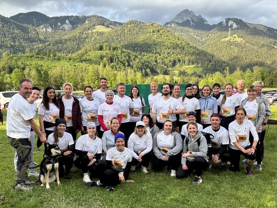 Eine große Gruppe von Menschen in weißen T-Shirts posiert lächelnd für ein Gruppenfoto auf einer Wiese vor einer Bergkulisse. Links im Vordergrund sitzt ein Hund.