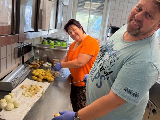 In einer Küche schälen ein Mann und eine Frau gemeinsam Kartoffeln. Beide tragen blaue Handschuhe und lächeln. Auf dem Edelstahltisch liegen große Mengen an Kartoffeln und Schalen, was auf eine ehrenamtliche Gemeinschaftskochaktion hindeutet.