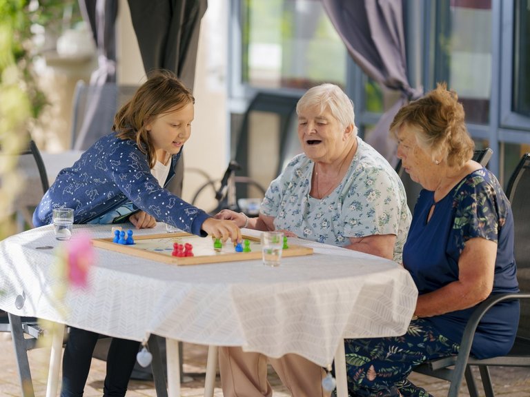 Ein Mädchen spielt Mensch Ärgere Dich nicht an einem Tisch mit zwei älteren Damen